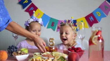 Bayram şapkası takan iki genç kız, meyve, fındık ve 5 mumlu sağlıklı bir doğum gününün tadını çıkarıyorlar. Birisi meyve tabağına mum koyarken heyecanlarını ifade ediyorlar.