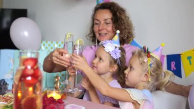 Anne sevinçle çocuklarının doğum gününü kutlar. Renkli süslemeler ve gülümseyen yüzlerle dolu neşeli bir kadeh kaldırıyorlar.