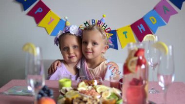Çiçekli iki kız kardeş, bir doğum günü partisi için dekore edilmiş bir masada, sağlıklı atıştırmalıklar, meyve suyu ve bir kutlama için hazırlanan boş bardaklarla kucaklaşıyorlar.