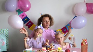 Mutlu bir aile, anne ve iki kız, doğum gününü renkli balonlar, parti şapkaları, süslü bir masa ve neşeli ifadelerle kutluyorlar. Sıcak ve neşeli bir ortam yaratıyorlar.