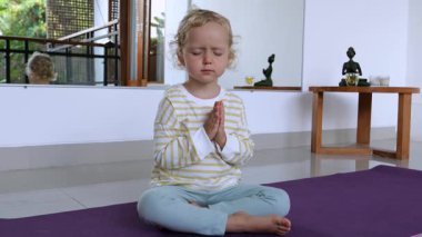 Küçük çocuk mor yoga minderi üzerinde nilüfer pozisyonu meditasyonu yapıyor. Huzurlu rahatlama tekniğini uygularken farkındalığı ve iç huzuru somutlaştırıyor.