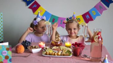 Doğum günü şapkası takan iki genç ikiz kardeş renkli pankartlar, mumlar, meyveler ve doğum günü tabelasıyla süslenmiş bir masada beşinci doğum günlerini kutluyorlar.
