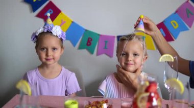 Ebeveynler parti şapkalarını ve saç bantlarını ikiz kızlarına takıyorlar, onları bir doğum günü kutlaması için hazırlıyorlar süslemeler ve içeceklerle.