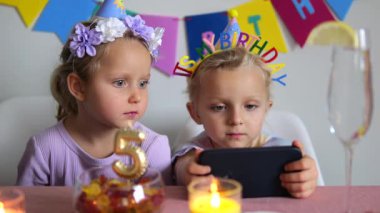 Doğum günü şapkası takan ikiz kardeşler yanan bir mum, bir bardak su, bir kase şeker ve arka planda mutlu bir doğum günü afişi olan bir masada oturmuş akıllı telefondan bir şey izliyorlar.