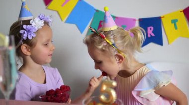 Parti şapkası takan iki genç kız beşinci doğum günü partisinde ahududu ve diğer yiyeceklerden zevk alarak neşeli ve kutlamalı bir ortam yaratıyorlar.