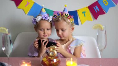 Parti şapkası ve elbisesi giyen iki genç kız, bir doğum günü süslemesi olan bir masada oturuyorlar, bir akıllı telefona bakıyorlar ve önünde mumlar ve bir kase şekerle gülümsüyorlar.