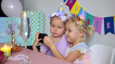 Renkli parti şapkaları takan iki kız kardeş doğum günü kutlaması sırasında akıllı telefon ekranlarını paylaşıyor dijital bağlantının ve birliktelik anının tadını çıkarıyorlar.