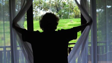 Arka ışıklandırılmış kadın gezgin otel odasındaki perdelerin arasından yemyeşil pirinç tarlalarını gösteriyor. Resimli ubud, bali manzarasında huzurlu bir neşe ve merak anı yaşıyor.