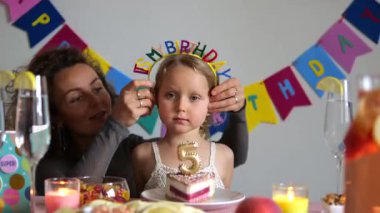 Neşeli bir ailenin doğum günü kutlaması. Anne sevgiyle kızların başına taç takıyor. Etrafı pasta, dekorasyon ve aile anlarıyla çevrili.