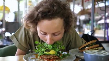 Sağlıklı ve lezzetli bir kahvaltının tadını çıkaran genç bir kadın üzerine yeşil sebze konmuş bir yemek, yanında da bir sandviç ve rahat bir kafede rahatlatıcı bir içecek.