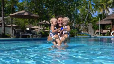 Mutlu anne, tropik bir tatil köyünde, palmiye ağaçlarıyla çevrili güzel bir mavi yüzme havuzunda oynayıp sıçrayan ikiz bebekleriyle vakit geçirmekten hoşlanıyor.