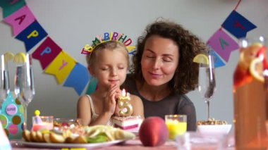 Doğum günü tacı takan sevimli bir kız, annesiyle bir şenlik masasında oturuyor, gözlerini kapıyor ve beşinci yaş günü pastasındaki mumları söndürmeden önce bir dilek diliyor.