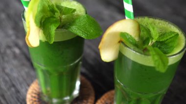 Taze nane yaprakları ve elma dilimleriyle süslenmiş iki bardak canlı yeşil detoks içeceği, sade bir ahşap masada oturup sağlıklı bir yaşam tarzını destekliyor.