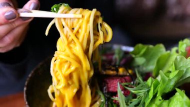 Ahşap çubuklar kullanan kadın eli, taze sebzelerle dolu renkli ramen kasesinden altın erişte kaldırma, iştah açıcı yemek sahneleri yaratan canlı malzemeler.