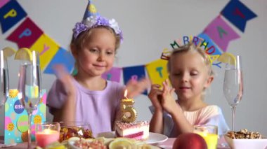 Şenlikli şapkalar takan iki genç kız el çırpıyor ve süslü bir pasta, meyve, mum ve masayı süsleyen renkli süslemelerle beşinci doğum günü partisini kutluyorlar.