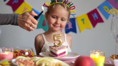 Beşinci yaş günü pastasına gülümseyen sevimli bir kız. Beş numaralı mumla süslenmiş, arka planda renkli bir afiş, masada mumlar ve meyveler var.