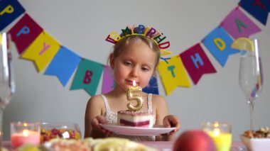 Doğum günü tacı takan, gözlerini kapayan ve doğum günü pastasındaki 5 numara şeklinde bir mum üfleyerek masasında renkli süslemeler ve abur cuburlar düzenleyen sevimli bir kız.