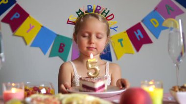 Sevimli kız beşinci yaş gününü kutluyor, bir dilek diliyor ve renkli pastasının üzerindeki mumu söndürmeye hazırlanıyor, etrafı şenlikli süslemeler ve ikramlar ile çevrili.