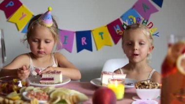 Parti şapkası takan ikiz kardeşler doğum günü partilerinde pasta yiyorlar ve arka planda renkli bir doğum günü afişi asılı.