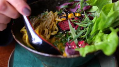 Canlı bir vejetaryen ramen kasesinden kırmızı kaşıkla çorba kepçeleyen bir kadına yakın durun. İçinde şehriyeler, renkli sebzeler ve bitki bazlı malzemeler olsun.