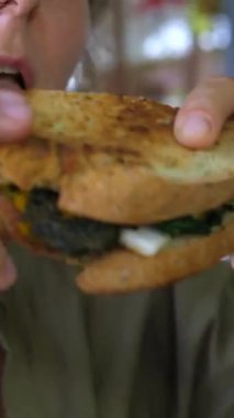 Dikey ekran genç kadın vejetaryen sandviçin tadını çıkartıyor. Keyifli bir öğle yemeği deneyimi yaşıyor. Günlük yemek mekanında taze, lezzetli bir yemeğin tadını çıkarıyor.