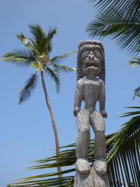 Hawaii Tanrısı 'nın oyulmuş heykeli. Güneş altında mavi gökyüzü arka planı ve palmiye ağaçları var.