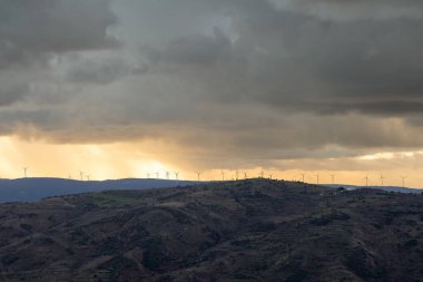 Uzak bir dağ sırtında, fırtına bulutlarını aşan günbatımı ışınlarıyla aydınlanan rüzgar türbinlerinin olduğu dramatik bir manzara. Çevresel yeşil elektrik 