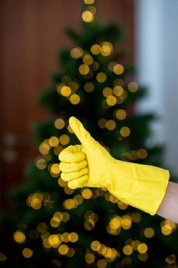 A person wearing yellow gloves sprays cleaning solution from a bottle, with a decorated Christmas tree and festive lights in the background. Professional cleaning  clipart