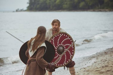 Hojbjerg, Danimarka, 27 Temmuz 2024: Viking çifti Viking festivalinde deniz kıyısında kılıçlarla savaşıyor.