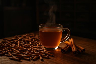 Yapay zeka, ahşap masadaki kurutulmuş ve taze tarçınla çevrili cam bardaktaki sıcak tarçın çayının görüntüsünü oluşturdu. Klasik Çin eczacılığı arka planı ve ılık düşük ışıklandırma atmosferi vardı.