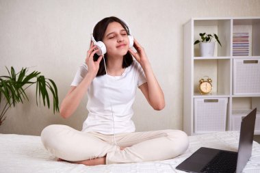 Kulaklıklı, gözleri kapalı müzik dinleyen genç bir kız. Kız rahat giysiler içinde odada yatağın üzerinde oturuyor. Model.