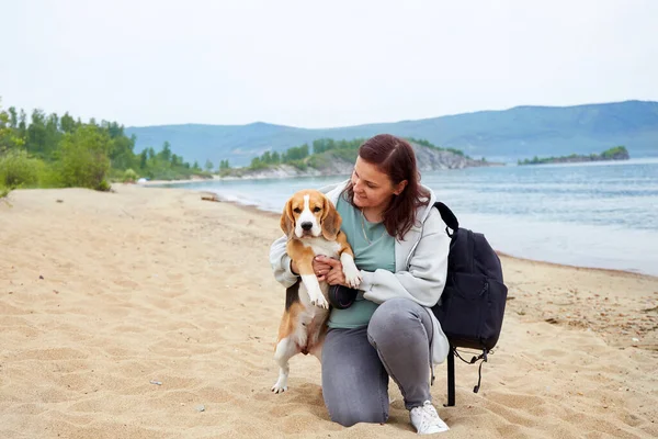 A woman traveler is sitting on a sandy beach in an embrace with her dog beagle. Summer holidays by the sea.