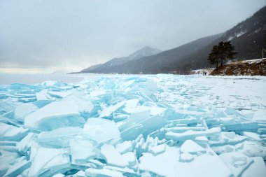 Donmuş buz blokları kıyı şeridini kaplayarak karla kaplı plajın yanında göz kamaştırıcı mavi bir manzara oluşturuyor. Arka planda dağlar yükseliyor ve kar yağıyor. Baykal Gölü 'nün kış manzarası. 