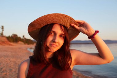 Gün batımında kumlu bir sahilde hasır şapkalı çekici bir kız. Kameraya bakıyorum..