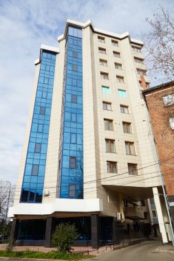 Şehir ortamında kocaman pencereleri olan modern çok katlı bir bina. Zemin katta yerleşim olmayan ticari binalar var..