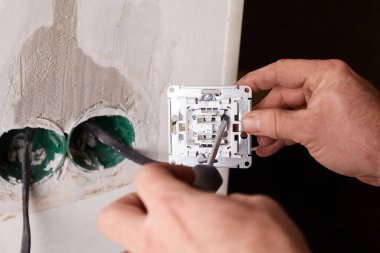 Bir işçi tornavida kullanarak duvara elektrik prizi takıyor. Arka planda, kısmen açık bir duvar yenileme çalışmalarını gösteriyor.