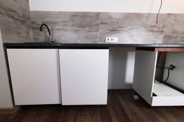Yeni yapılmış mutfak tezgahında, yenilenmekte olan bir dairede dokulu gri bir duvara dayalı siyah bir lavabo ve beyaz bir dolap yer alıyor.. 