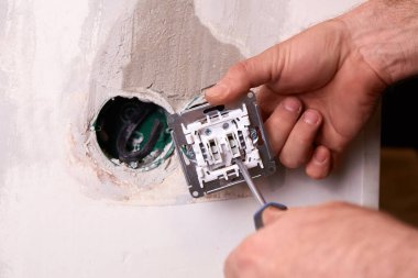 Bir işçi tornavida kullanarak duvara elektrik prizi takıyor. Daire tadilattan geçiyor..