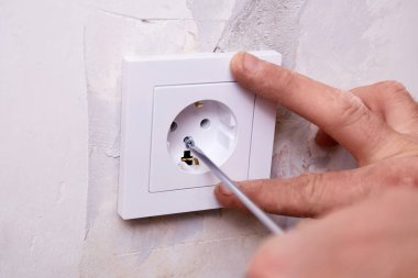 Bir kişi duvara elektrik prizi takmak için tornavida kullanıyor. Duvar yenileme çalışmalarının devam ettiğini gösteriyor..
