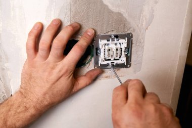 Bir işçi tornavida kullanarak duvara elektrik prizi takıyor. Daire tadilattan geçiyor..