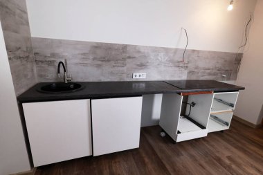 Yeni yapılmış mutfak tezgahında, yenilenmekte olan bir dairede dokulu gri bir duvara dayalı siyah bir lavabo ve beyaz bir dolap yer alıyor.. 