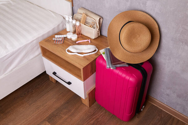 Hotel room interior. Personal hygiene items, travel cosmetics on the bedside table, bright pink suitcase with a straw hat. Travel concept