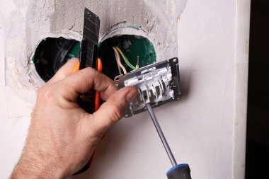 Bir işçi tornavida kullanarak duvara elektrik prizi takıyor. Daire tadilattan geçiyor..