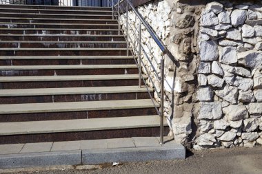 Gri betondan yapılmış ve metal parmaklıklarla süslenmiş katı bir merdiven engebeli bir taş duvara dayanır. Güneş ışığı sağlam tasarımı vurguluyor, ziyaretçileri davet ediyor. 