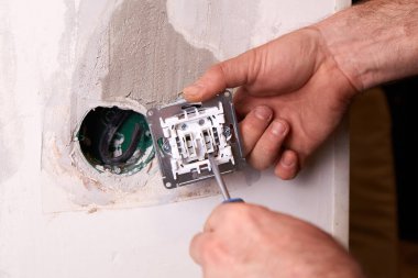 Elektrikçi beyaz bir duvara elektrik prizi takmak için tornavida kullanıyor. Çevre minimalistik, bu da yeni bir ev yenileme projesine işaret ediyor.. 