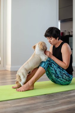 Esmer, orta yaşlı bir kadın evde köpekle egzersiz minderi üzerinde yoga yapıyor, akıl sağlığı, zihinsel denge, kendine zaman ayırıyor, stres atma, arabuluculuk, zihin dolgunluğu konsepti.