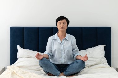 Esmer, orta yaşlı, gömlek ve kot pantolonlu bir kadın yatak odasında yoga meditasyonu yapıyor, akıl sağlığı yerinde. Huzurlu kadın zihinsel dengeyi sağlamak için meditasyon yapar, evde kendine zaman ayırır..