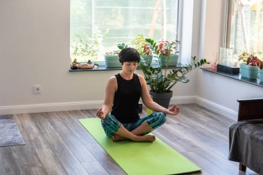 Esmer, orta yaşlı bir kadın evde egzersiz paspası üzerinde yoga meditasyonu yapıyor, akıl sağlığı yerinde. Huzurlu kadın meditasyon, zihinsel denge, kendine zaman ayırmak, stres atma, arabuluculuk, zihin doluluk konsepti.
