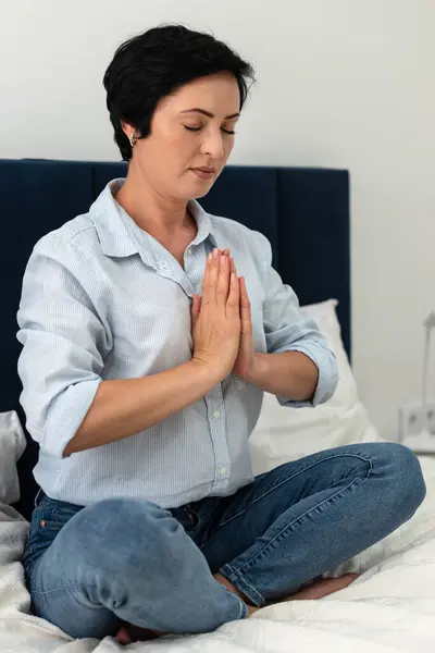 Brunette middle aged woman in a shirt and jeans doing yoga meditation at home in the bedroom, mental health. Peaceful woman meditate to achieve mental balance, time for herself at home.