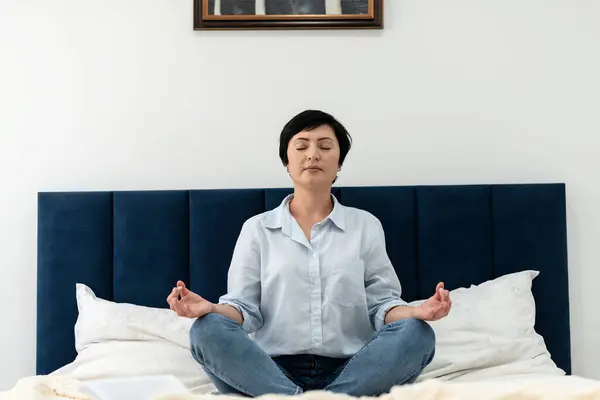 Brunette middle aged woman in a shirt and jeans doing yoga meditation at home in the bedroom, mental health. Peaceful woman meditate to achieve mental balance, time for herself at home.
